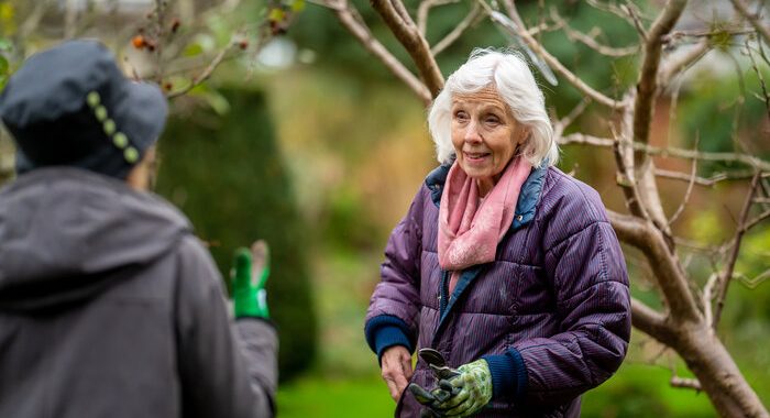 Two older women in a garden with trees. One is looking at the other and they both have gardening gloves on