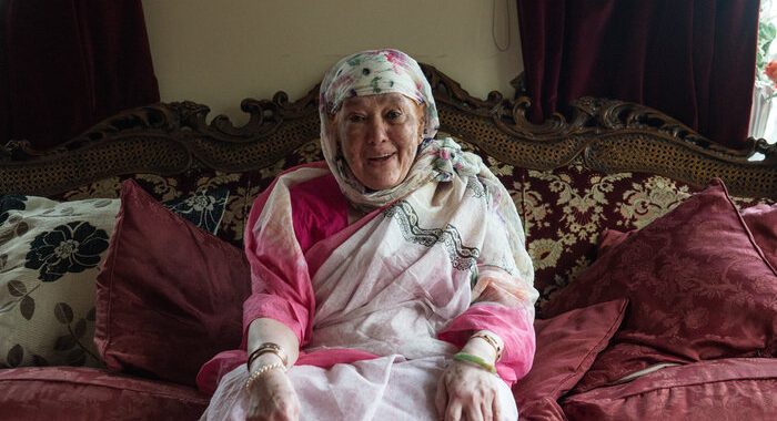 An older lady sitting at home on the sofa. She's wearing a colourful headscarf