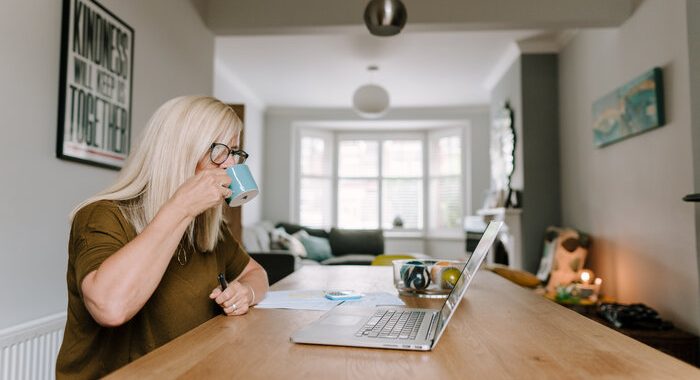 Woman drinking out of a mug, sitting in her house and looking at a laptop