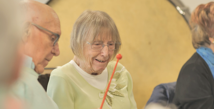 An older woman smiling and holding a drumstick with an older man sitting next to her