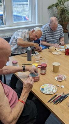 Three older man sitting next to each other at a table and decorating clay pots