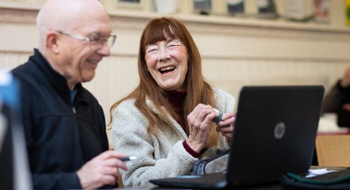 An older woman and older man sitting at a table with a laptop. The man is smiling and looking at the laptop and the woman is looking at the man and laughing