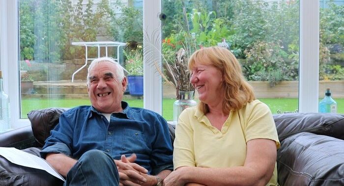 Photo of Liz and Mike sitting and smiling in front of a large window with their garden behind them. Liz and Mike are members of our Dementia Carers Expert Reference Group