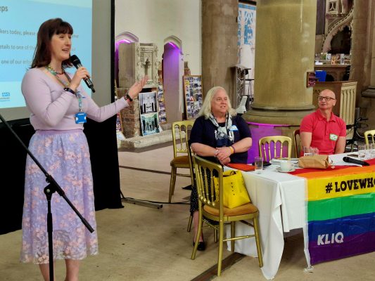 Sarah Fox talking into a microphone with Daithi Clayton and John Hammond sitting behind a table facing the audience