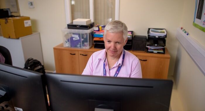 A woman at work sitting behind a desk in an office. She is looking at two computer screens