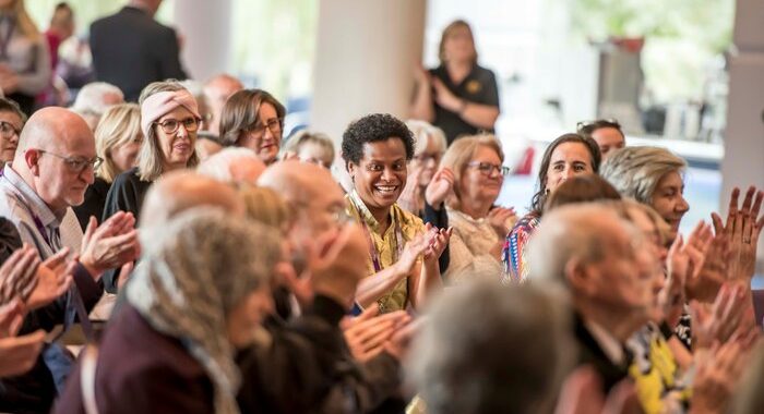 A group of people in a large hall sitting as an audience, People are clapping and some are smiling