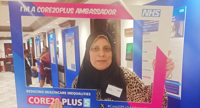 Amal Morsi, Project Manager with Dementia United standing behind a frame. The frame reads I'm a Core20Plus Ambassador. The NHS logo is in the top right hand corner of the frame and at the bottom left of the frame it reads - Reducing Healthcare Inequalities Core20Plus5