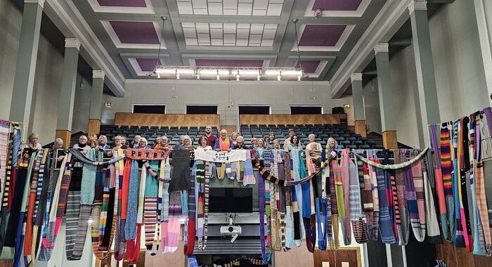 Lewy Body Society ambassador Christopher Biggins, Scarf For Lewy founder Vicky Hands, Lewy Body Society CEO Jacqui Cannon and some of the mile of scarves knitted for Lewy Body Society’s Scarf for Lewy at Manchester’s historic Central Hall. They are standing on a balcony with lots of scarves draped over the edge and there are many of the knitters standing behind them