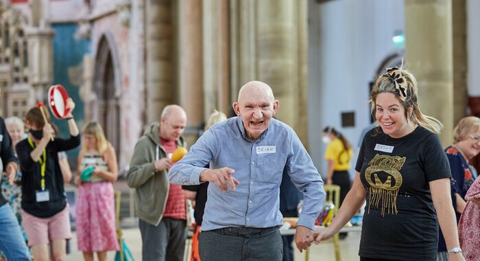 A photo taken at Gorton Monastery. There are a lot of pole. One has a tambourine, another a maraca. At the front of the photo there's a young woman facing the camera and smiling. She's holding the hand of an older man and he is facing the camera and smiling