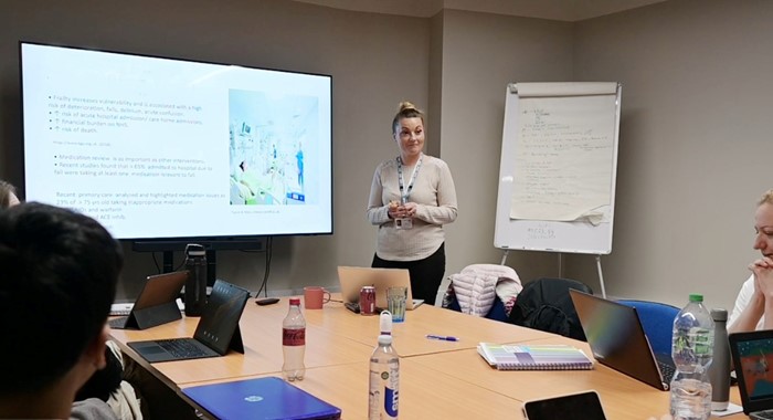 A photograph showing a group of people sitting round a table with laptops. At the front of the room there's a woman standing in front of a screen showing a presentation